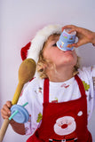 Child in a Santa hat and apron holding a wooden spoon and drinking from a cup with a white background