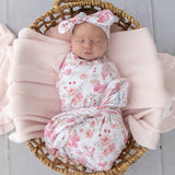 A sleeping newborn baby is swaddled in a soft pink and white floral blanket and wearing a matching bow headband, resting inside a woven wicker basket lined with a pale pink knit blanket.