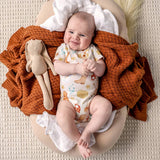 A baby lying on a beige cushion, wearing a white bodysuit with a colorful boot print, surrounded by an orange textured blanket and a beige stuffed bunny.
