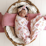 A sleeping newborn baby swaddled in a white muslin blanket with pink and gold baby boot prints, wearing a matching pink and gold printed headband, resting inside a woven wicker basket with a pink knit pillow.