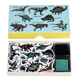A dinosaur-themed stamp set shown open with its lid placed above. The lid is light blue with black dinosaur illustrations and labels.