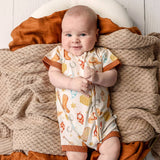A baby lying on a textured beige blanket and rust-colored pillows, wearing a short-sleeve one-piece romper with a white base and a colorful western-themed print including boots, horseshoes, and stars.