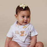 A baby wearing a white short-sleeve bodysuit and matching bib with a cute watercolor animal print, sitting against a solid beige background. The baby has a pale yellow bow in their hair.