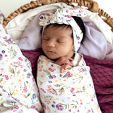 A sleeping newborn baby swaddled in a white floral blanket and wearing a matching floral bow headband, resting inside a woven basket with soft blankets.
