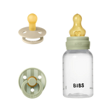 BIBS baby bottle and pacifier set in sage green and pale yellow, shown on a clean white background. The set includes a clear bottle with a pale yellow teat, a sage green collar, and a black BIBS logo, alongside two pacifiers (one beige and one striped in sage green and pale yellow).