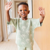 Child wearing a light green sweater with palm tree patterns, standing indoors.