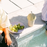 Person sitting by a pool with grapes on a plate and a beige cup.