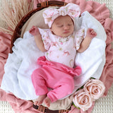 A sleeping newborn baby wearing a light pink floral romper, bright pink ruffled pants, and a matching pink bow headband, lying in a wicker basket lined with white fabric and surrounded by pink textiles and faux roses.