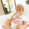 Baby sitting on a white chair holding a purple teething toy with palm trees in the background