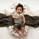 Baby lying on a cushion with dinosaur pattern, wearing a striped shirt and green shorts.