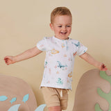 A young toddler boy wearing a white short-sleeve t-shirt with a colorful sea-creature print and beige shorts, standing in a playful studio setting with decorative props.