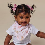 A smiling toddler with dark hair in two pink bows wears a white ribbed bodysuit and a light pink patterned bib. The bib features illustrations of ballet slippers, a bow and arrow, and a horseshoe.