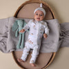 Baby in a white onesie with animal patterns lying on a textured blanket next to a green plush toy.