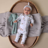 Baby in a white onesie with animal patterns lying on a textured blanket next to a green plush toy.