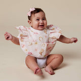 A baby sitting on a beige studio floor wearing a ruffled bib with a pink and gold western-themed print, including boots, horseshoes, and flowers. The baby is also wearing a pink bow in her hair.