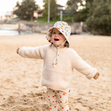 Child wearing a colorful sun hat and beige sweater on a sandy beach.