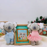 Two plush koalas, one in overalls and one in a dress, standing next to a framed picture of two koalas.