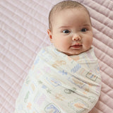 A baby swaddled in a soft, cream-colored muslin wrap with pastel multicolor sea life and geometric patterns, lying on a pink quilted mattress.