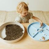 Child playing with a blue tray and a white bowl filled with sand on a wooden surface.
