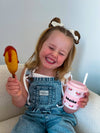 Child holding a toy corn dog and a pink cup with a straw on a light blue background