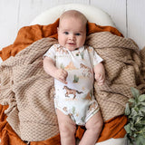 A baby lying on a white wooden floor, wrapped in a textured beige blanket and an orange throw, wearing a white short-sleeve bodysuit with a desert safari print of cacti, camels, and horses.
