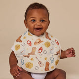 A baby wearing a white bib with a western-themed print of cowboy boots, horseshoes, hats, and stars, photographed against a solid beige background.