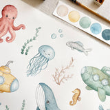 Watercolor underwater illustration printed on white paper, featuring an octopus, jellyfish, fish, submarine, whale, seahorse, and coral, with a paint palette and pencil visible at the top right.