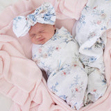 Newborn baby wrapped in floral fabric with a matching headband, lying on pink textured fabric.