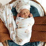 Newborn baby swaddled in a blanket with hot air balloon pattern, wearing a matching hat, in a woven basket.