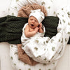 Newborn baby wrapped in a swaddle with palm tree pattern, wearing a white hat, on a textured surface.