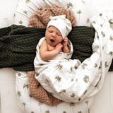 Newborn baby wrapped in a swaddle with palm tree pattern, wearing a white hat, on a textured surface.