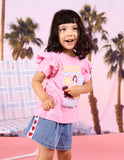 A young girl with dark hair smiling and posing in a pink graphic t-shirt with ruffled sleeves and a denim skirt, set against a pink tropical beach backdrop with palm trees.