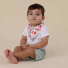 Baby sitting on a beige background wearing a white shirt with a floral bib and light green shorts.