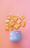 Small blue container with cartoon designs on a pink background, surrounded by crackers and popcorn.