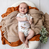 A baby lying on a white cushion, wearing a white short-sleeve bodysuit with a safari animal print. The baby is surrounded by a chunky beige knit blanket and an orange rust-colored fabric, with a small green plant in the corner.