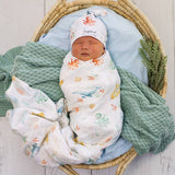 A newborn baby is swaddled in a white muslin blanket with colorful sea-creature prints, wearing a matching white hat. The baby is lying in a woven rattan bassinet, surrounded by a light blue cushion, a green knit blanket, and a small green plant.