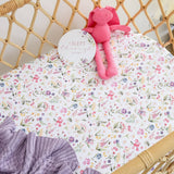 A close-up of a baby crib bedding set featuring a white fitted sheet with a colorful floral print and a lavender knit blanket, styled in a rattan crib with a pink plush toy and a round tag that reads 'I SLEPT through the night'.
