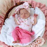 A newborn baby wearing a pink floral short-sleeve romper and matching pink pants, with a large bow headband, lying on a white blanket inside a wicker basket with pink fabric and dried grass around them.