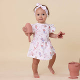 Baby in a floral dress holding a pink watering can on a beige background