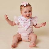 A baby sitting on a beige studio background wearing a pale pink short-sleeve romper, a white bib with a pink flamingo print, and a matching pink bow in her hair.