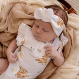 A sleeping newborn baby wearing a white short-sleeve romper and matching bow headband, both printed with cute brown and yellow ducklings and small green leaves, wrapped in a beige blanket.