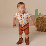 A toddler stands in a studio setting wearing a white short-sleeve top with a cowboy-themed print, rust brown shorts, and brown cowboy boots. The background features a beige backdrop, a decorative green cactus, and a bale of hay.