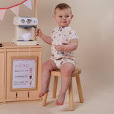 A baby wearing a cream-colored short-sleeve romper with a small multicolor food pattern sits on a wooden stool next to a toy play kitchen with a mini espresso machine.
