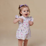 A toddler girl with a purple bow in her hair is wearing a white short-sleeve romper with a colorful floral print, standing against a solid beige studio background.