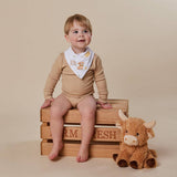 A baby wearing a beige long-sleeve bodysuit and a white printed bandana bib sits on a wooden crate next to a plush brown cow toy, against a solid beige studio background.