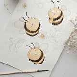 A watercolor-style illustration of three cute bees on a white sheet of paper, with floral line drawings and a paintbrush nearby. The bees are cream, yellow, and black, and one holds a honey pot.
