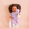 Doll in purple floral pajamas holding a baby bottle and rattle on a pink background