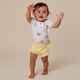 A toddler standing barefoot on a plain beige studio background, wearing a white short-sleeve top with a light brown animal print and pale yellow ruffled shorts.