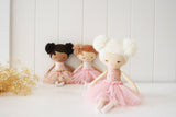 Three dolls in pink dresses sitting on a white surface with a light wooden panel background.