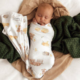 A sleeping newborn baby is swaddled in a white muslin swaddle blanket with a brown and yellow woodland animal print, styled on a tan sheet with a dark green chunky knit blanket.